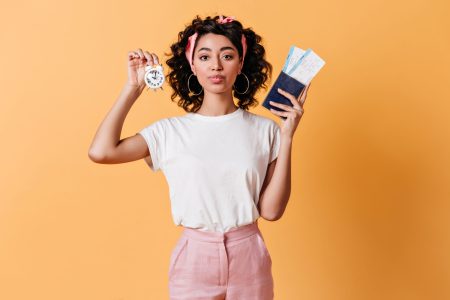 Beautiful girl holding tickets and alarm clock. Front view of stylish woman with passport posing on yellow background.